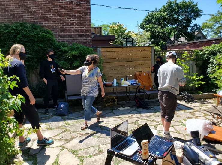 Several artist form a distanced circle outside. Jacquie Thomas stands in the middle with an audio recording device in hand outstretched towards the artists.